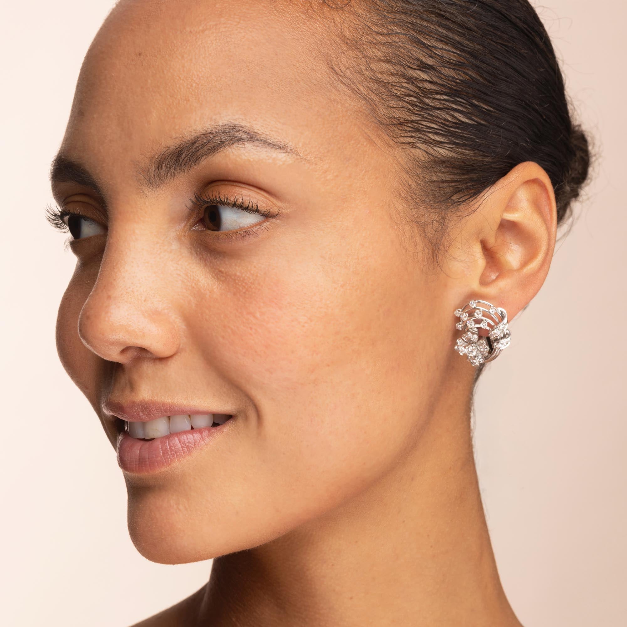 A close-up side profile of a woman wearing a silver-toned, flower-like cluster stud earring with clear stones against a soft beige background.