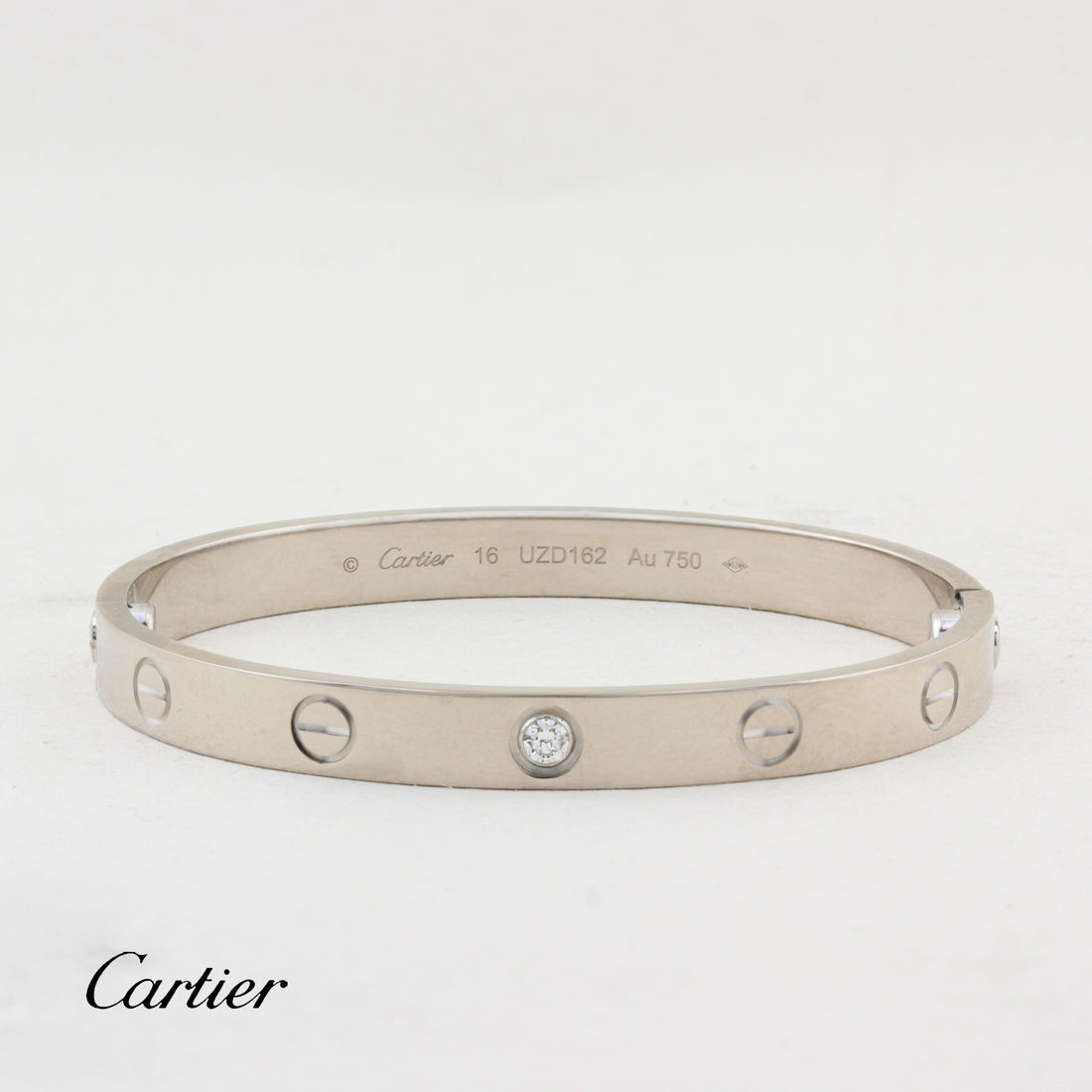 A Cartier Love bracelet in white-tone metal with five screw details and a central clear stone, shown on a white background with the Cartier logo at the bottom left.