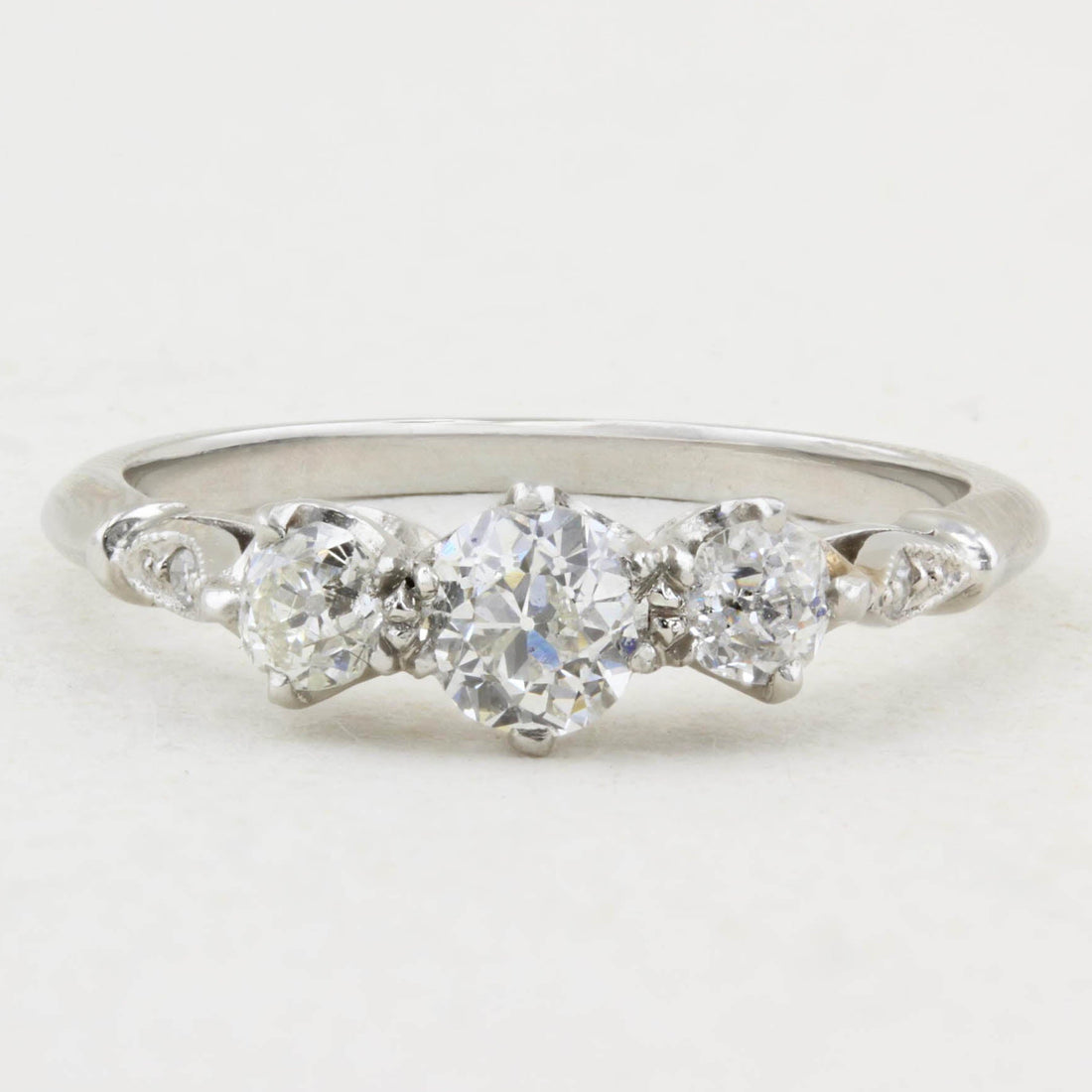 A white gold three-stone diamond ring with a central round diamond flanked by two smaller round diamonds, accented by small diamonds on the shoulders, set against a plain white background.