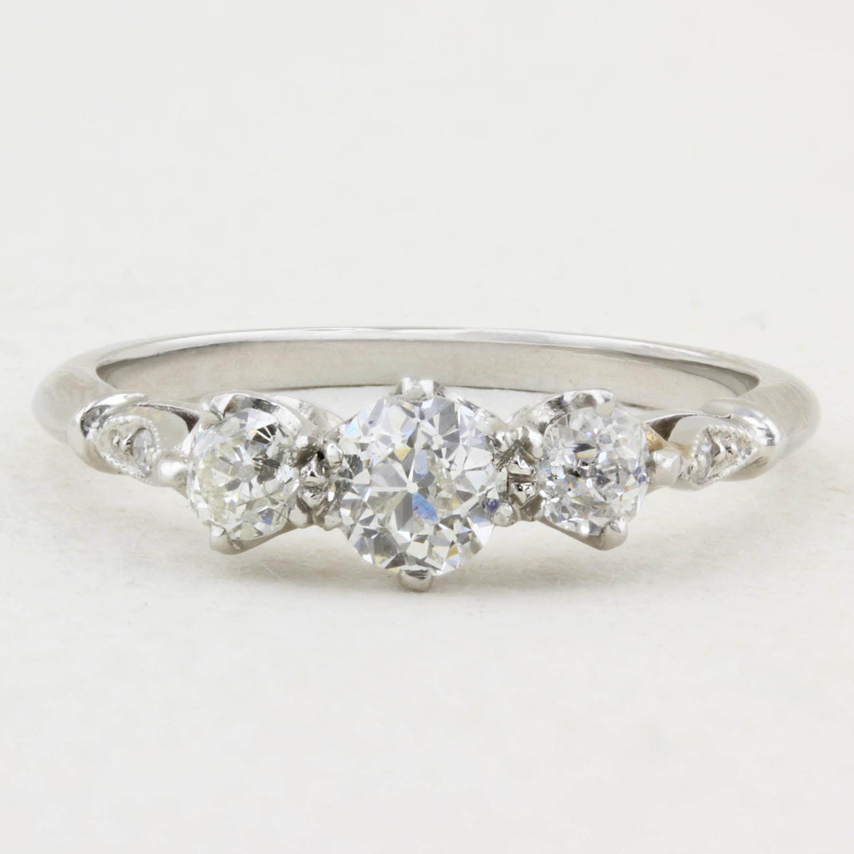 A white gold three-stone diamond ring with a central round diamond flanked by two smaller round diamonds, accented by small diamonds on the shoulders, set against a plain white background.