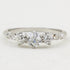 A white gold three-stone diamond ring with a central round diamond flanked by two smaller round diamonds, accented by small diamonds on the shoulders, set against a plain white background.